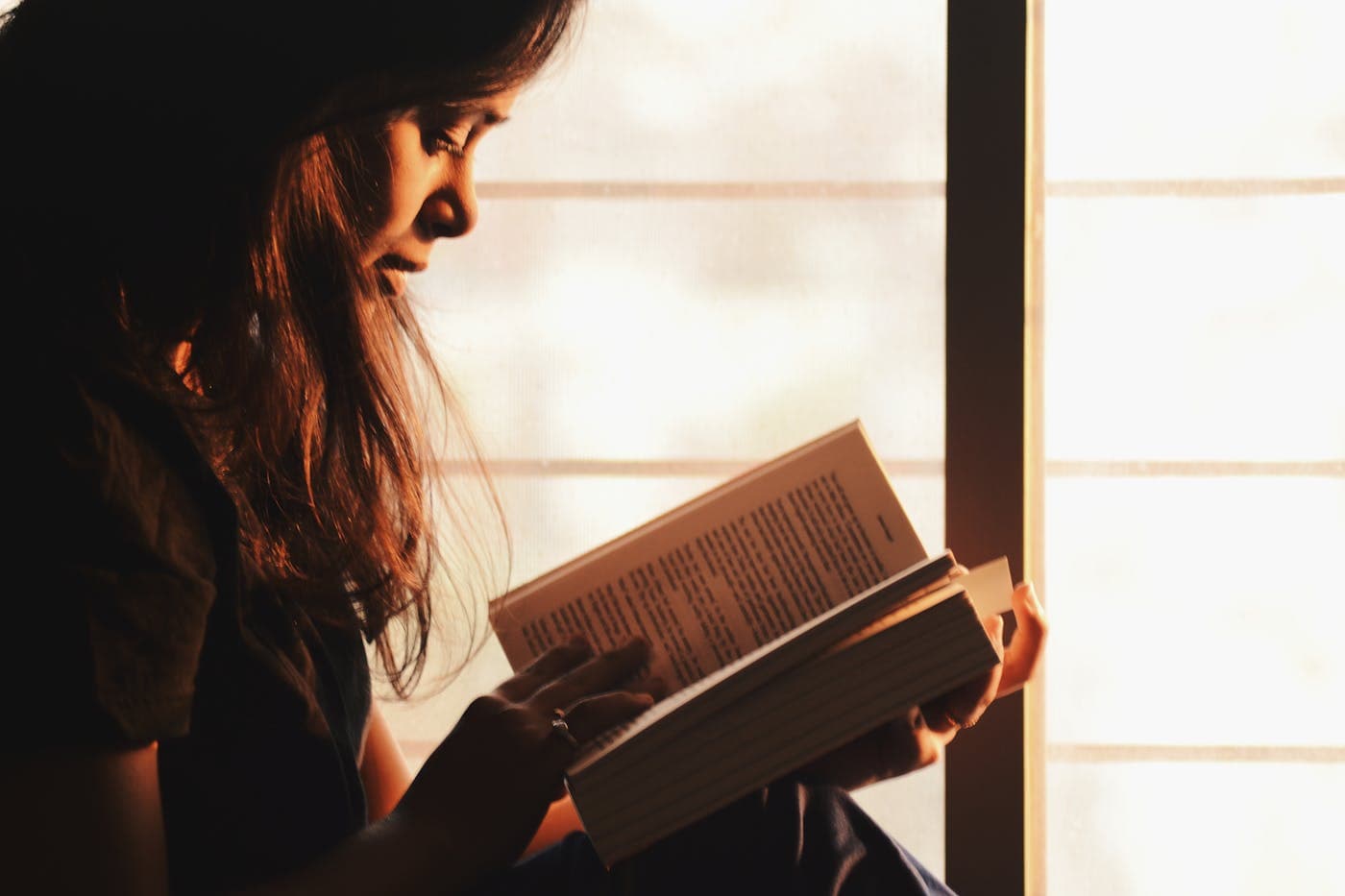 A person reading a book beside a sunlit window.