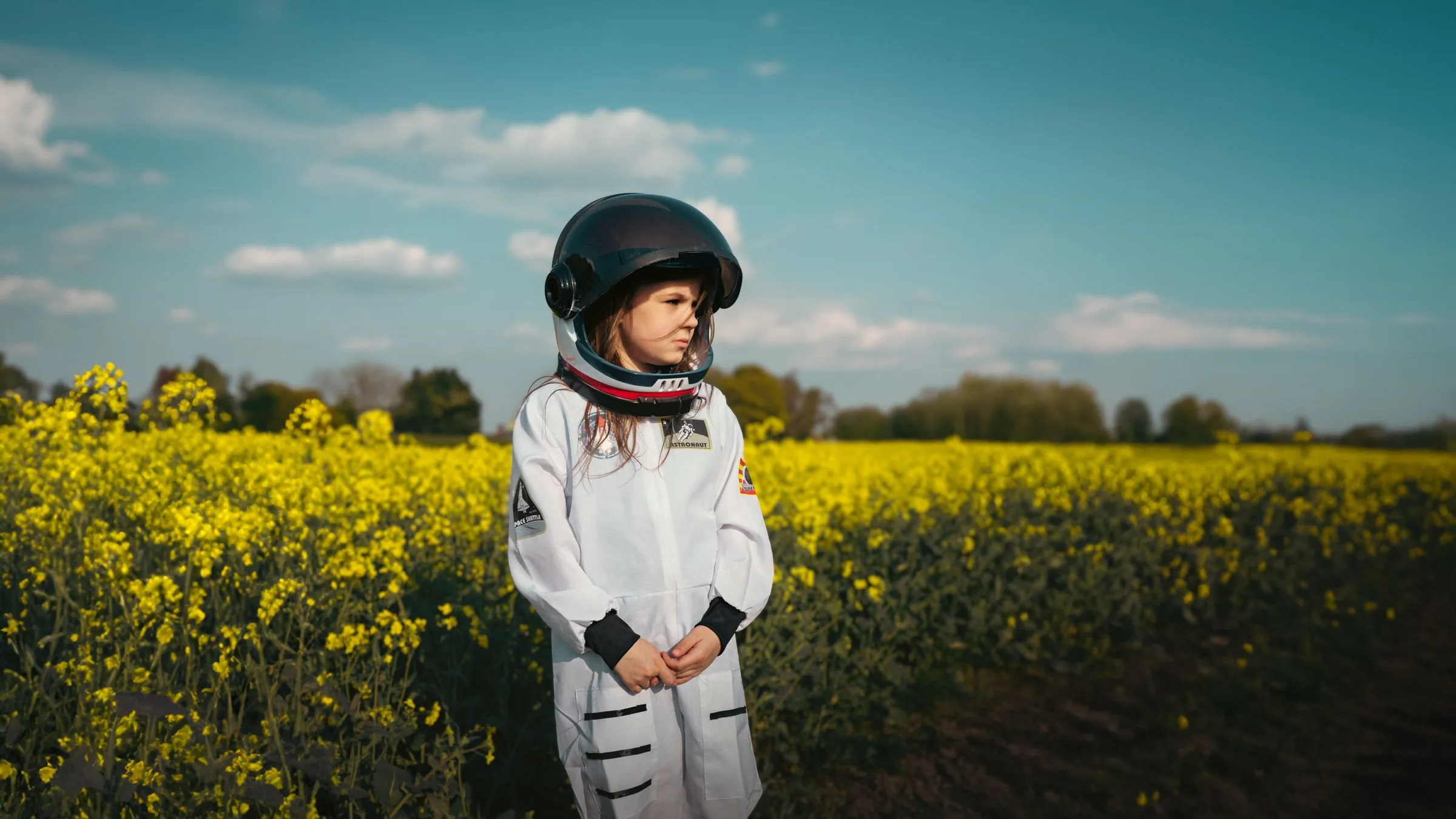 A small girl in a white astronaut suit and helmet standing in a field of yellow flowers under a blue sky