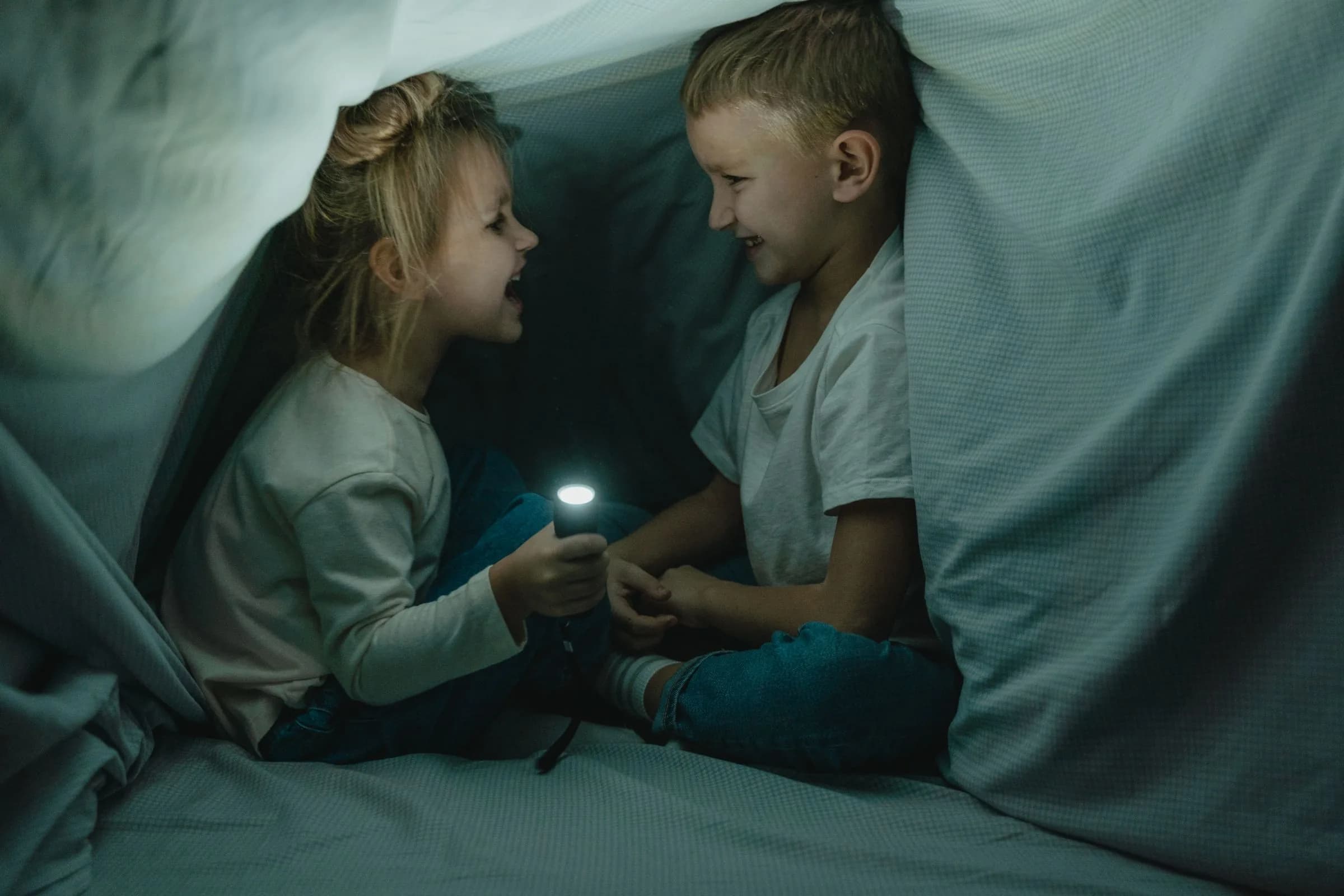 Two children laughing inside a blanket fort, lit by a flashlight