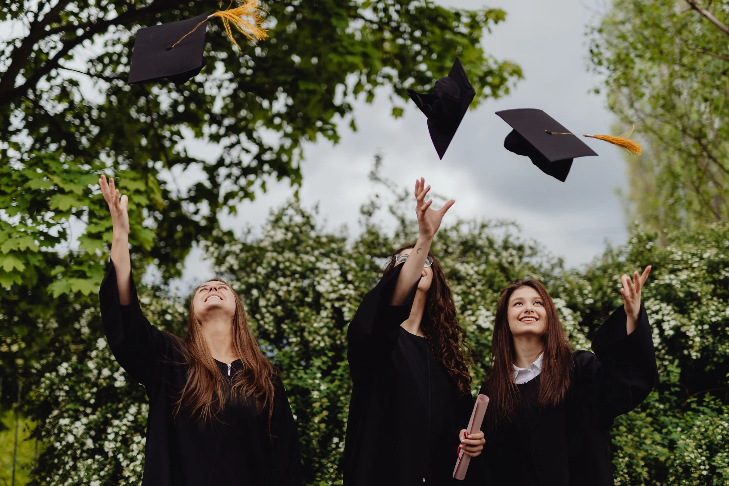 Three graduates in gowns throwing their caps into the air under spring trees
