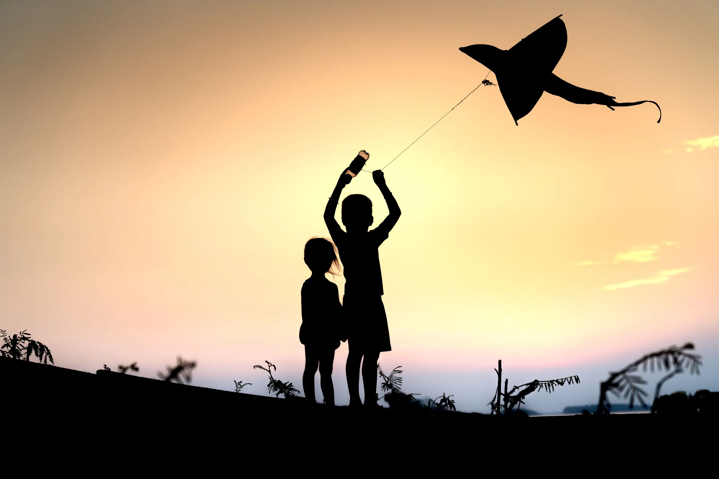 Two children in silhouette flying a kite against a sunset sky