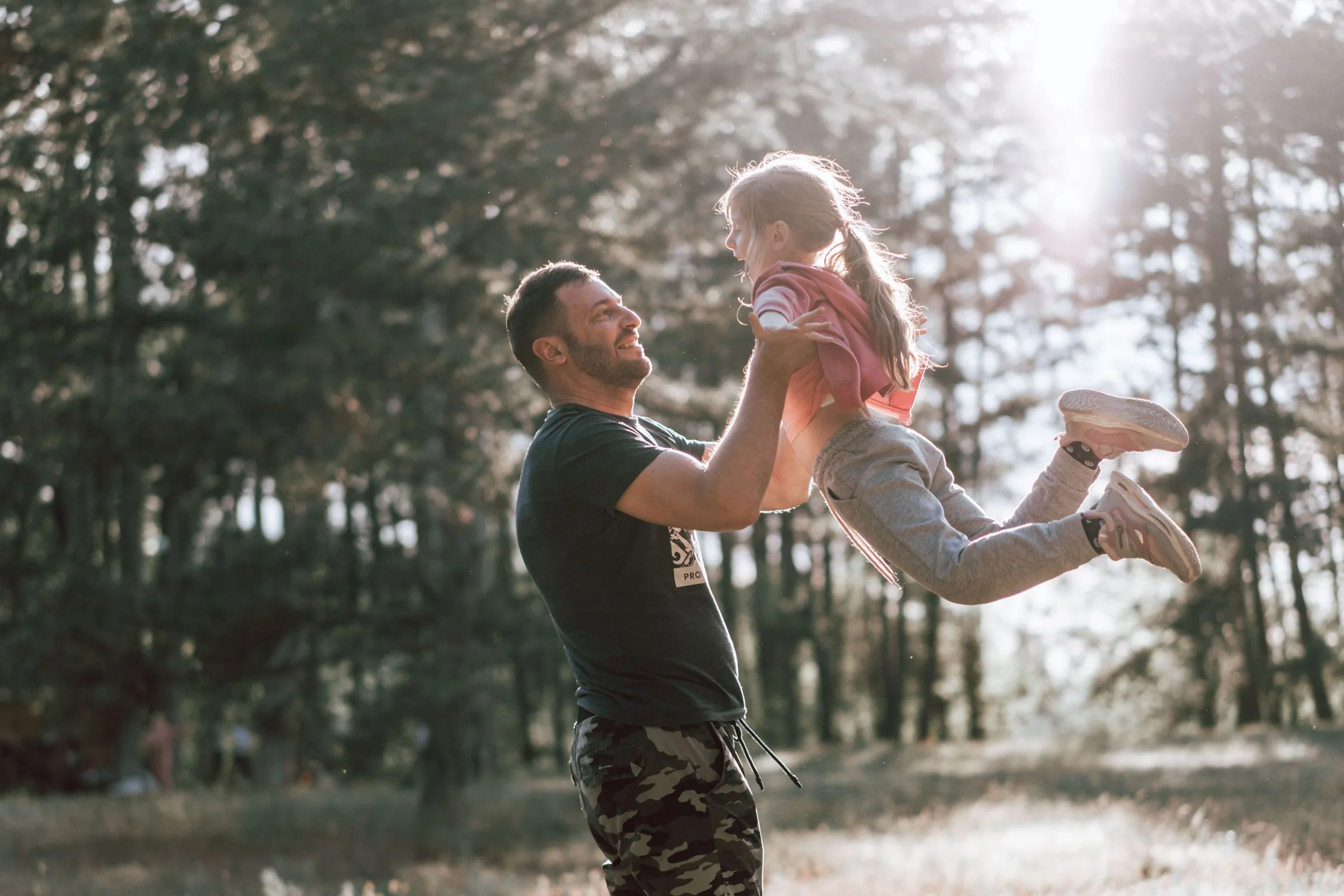 A father lifting his laughing daughter into the air in a sunlit pine wood