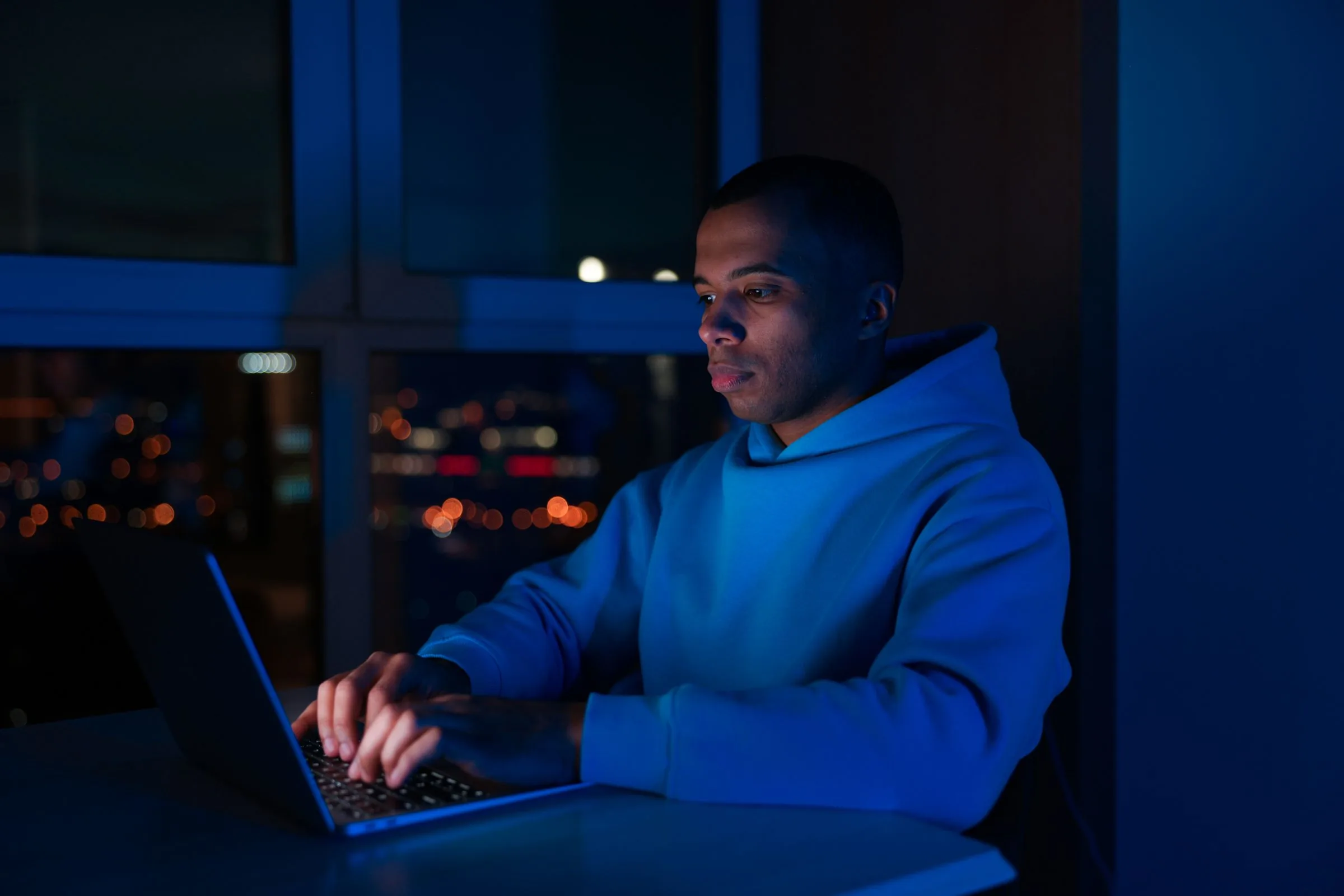 A man typing on a laptop at night, lit blue by the screen, city lights behind him