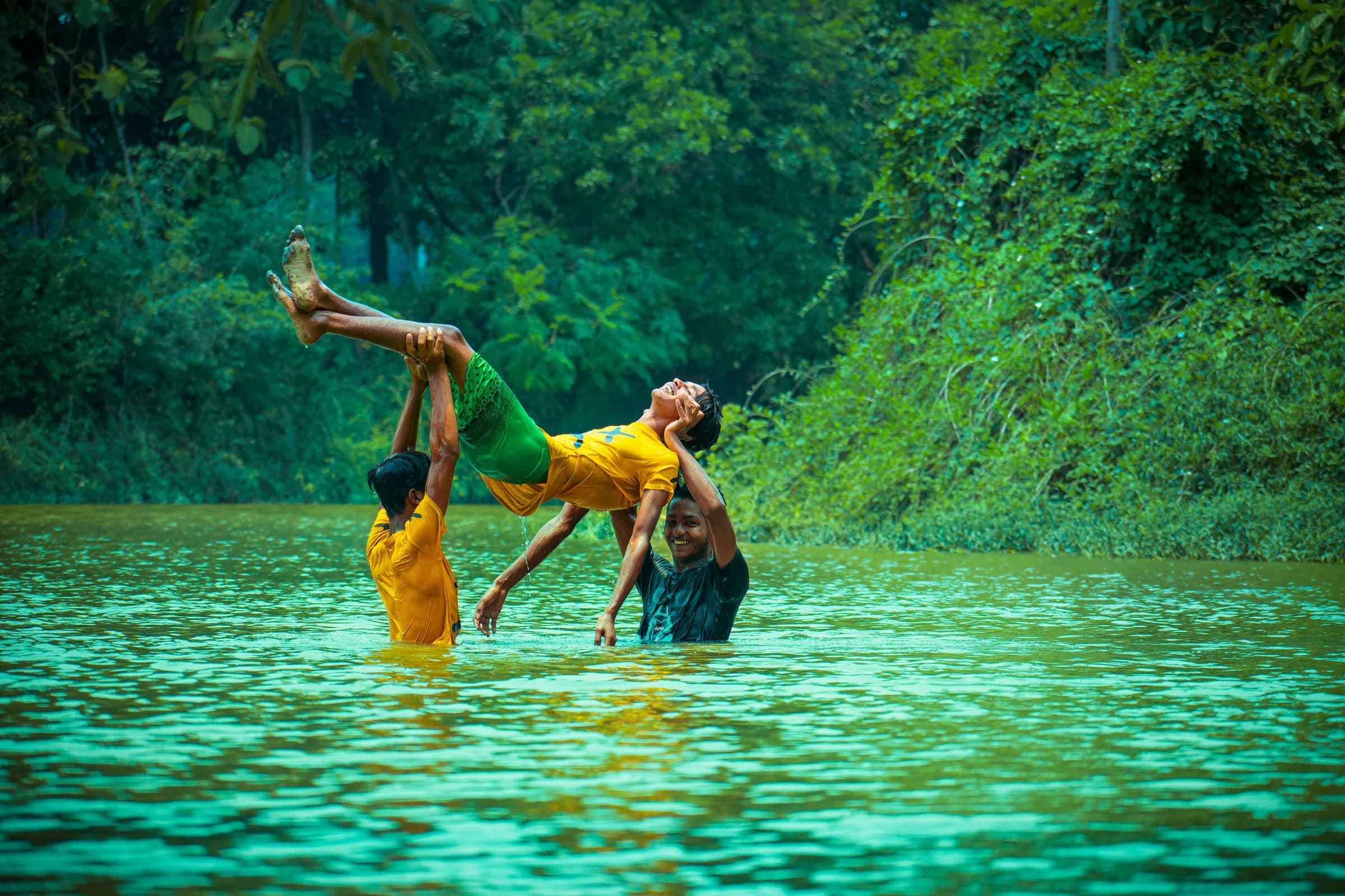 Three friends playing in a green river, two of them tossing the third into the air
