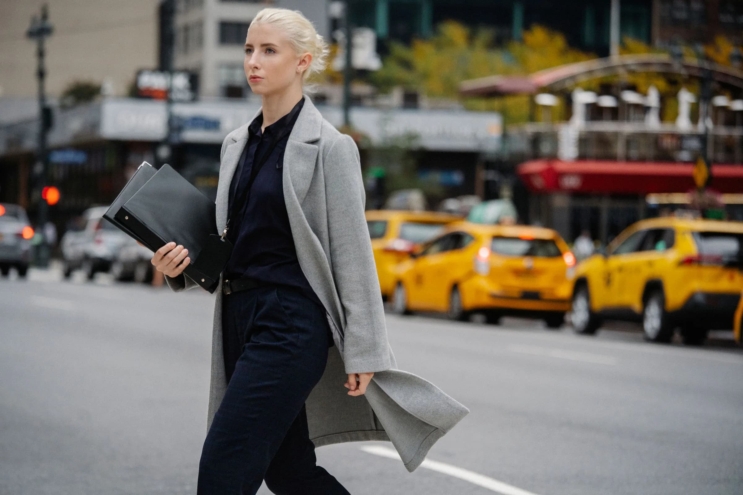 A woman in a long grey coat striding across a city street, folders in hand, yellow taxis behind her