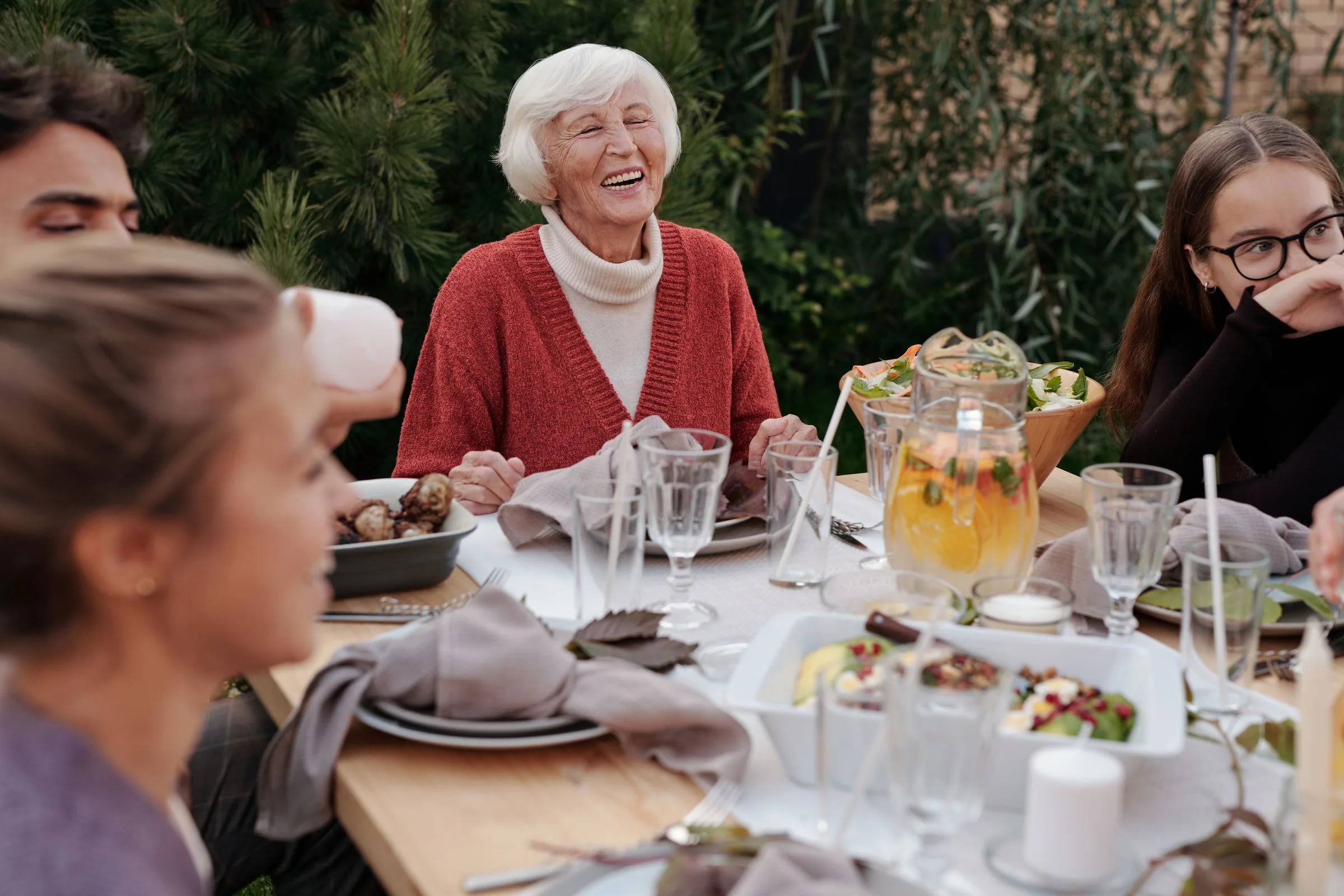 A grandmother laughing at a long garden table set for dinner, family and friends around her