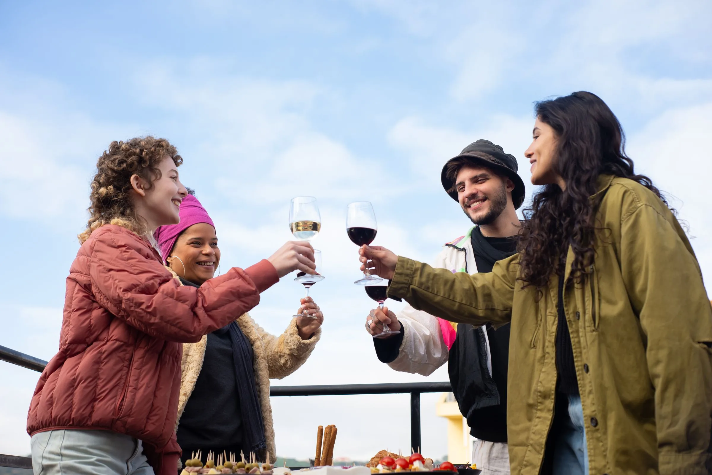 Four friends raising wine glasses across a rooftop table under a blue sky