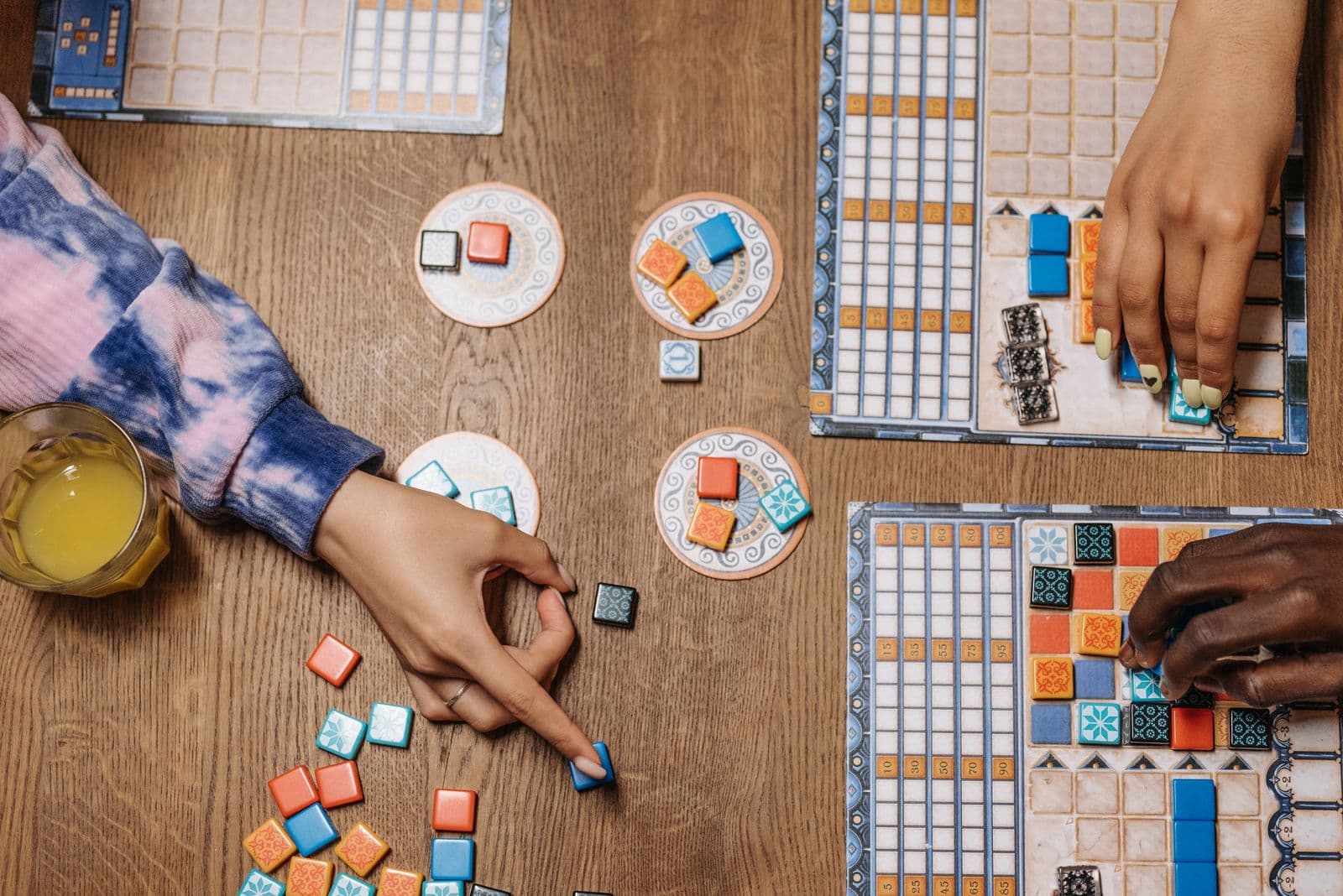 Players arrange colorful tiles on board games around a wooden table.