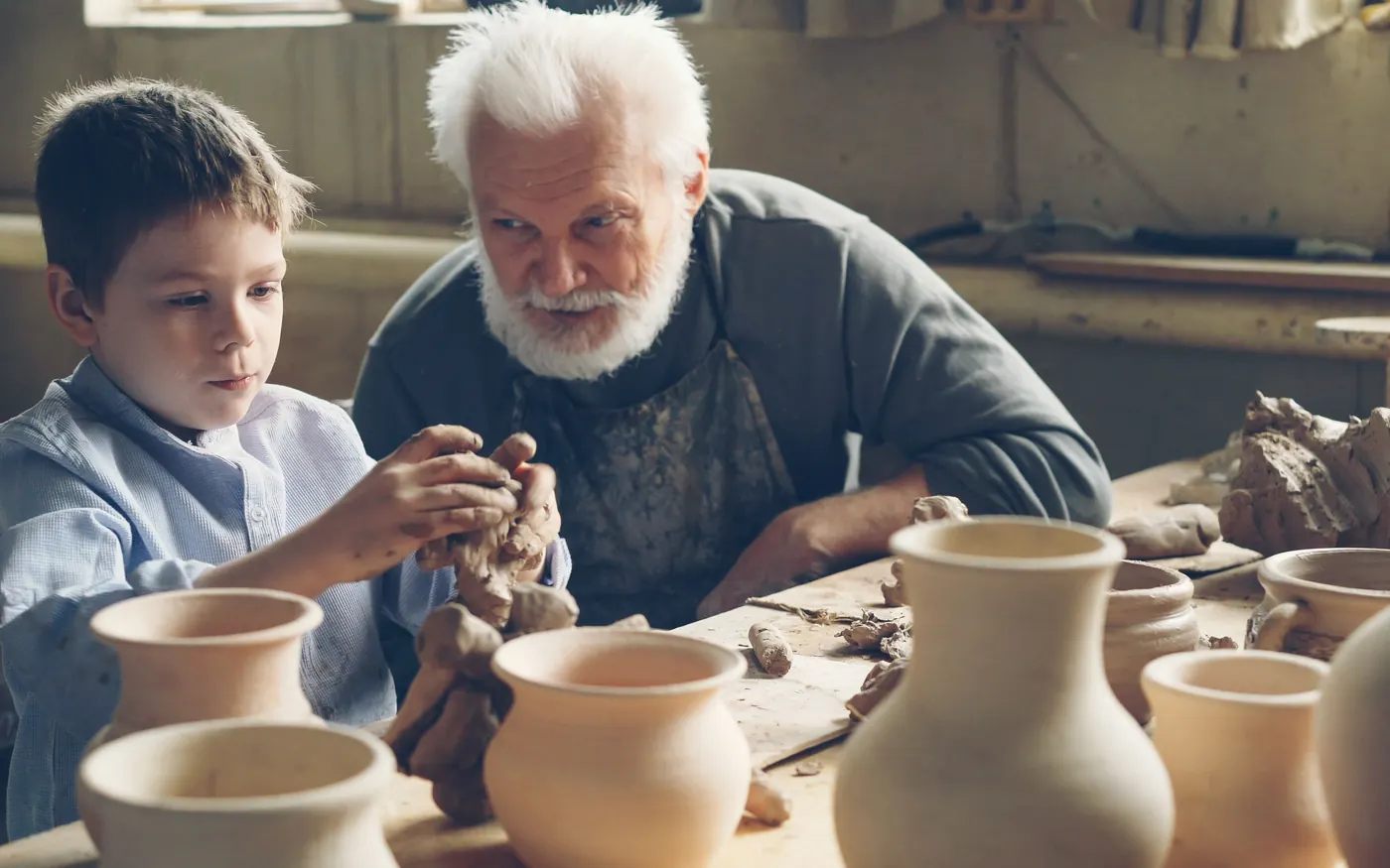 A grandfather and grandson shaping clay together in a pottery workshop