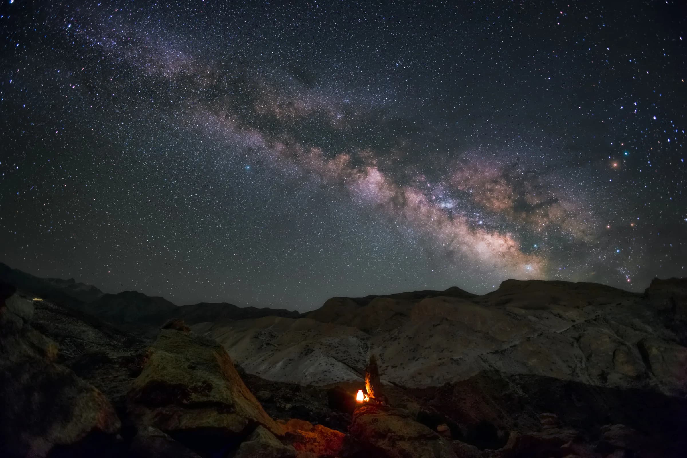 The Milky Way arching over a dark desert canyon, a small campfire glowing among the rocks below