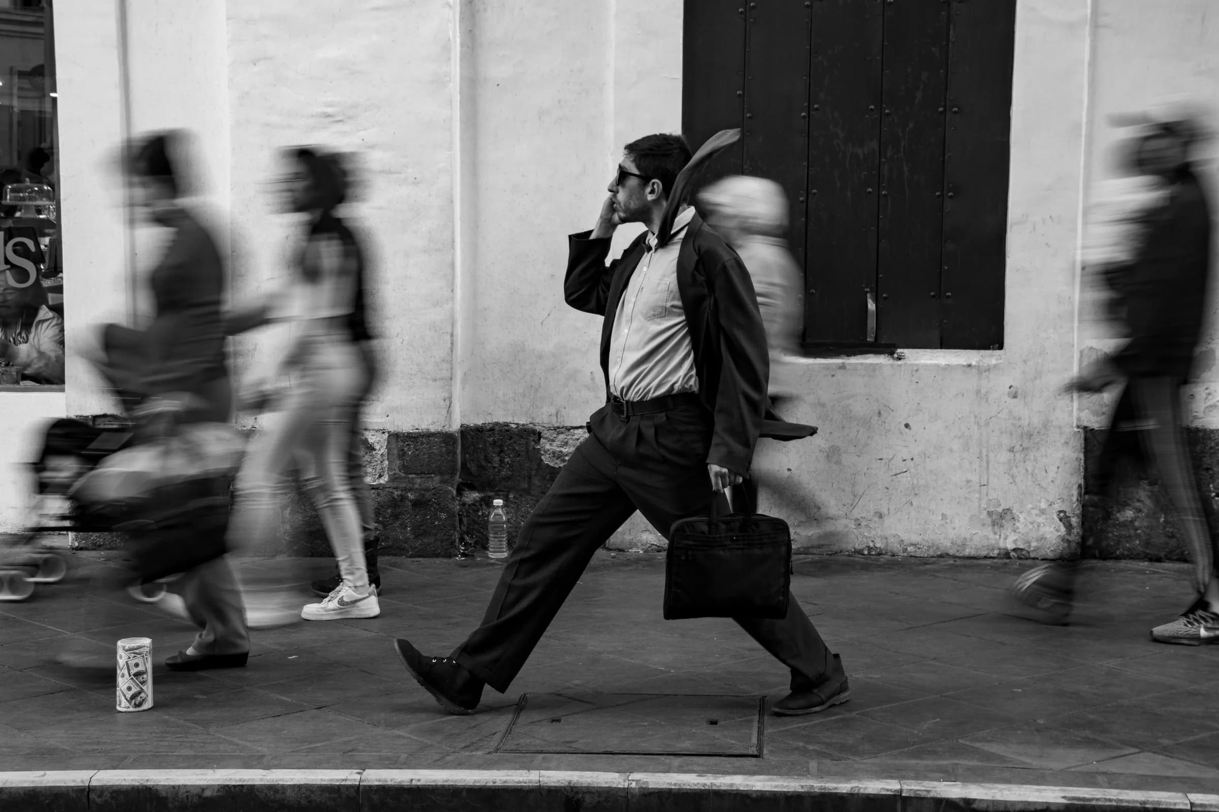 A man in a suit striding along a city pavement with a phone to his ear and a briefcase in hand, passers-by blurred around him