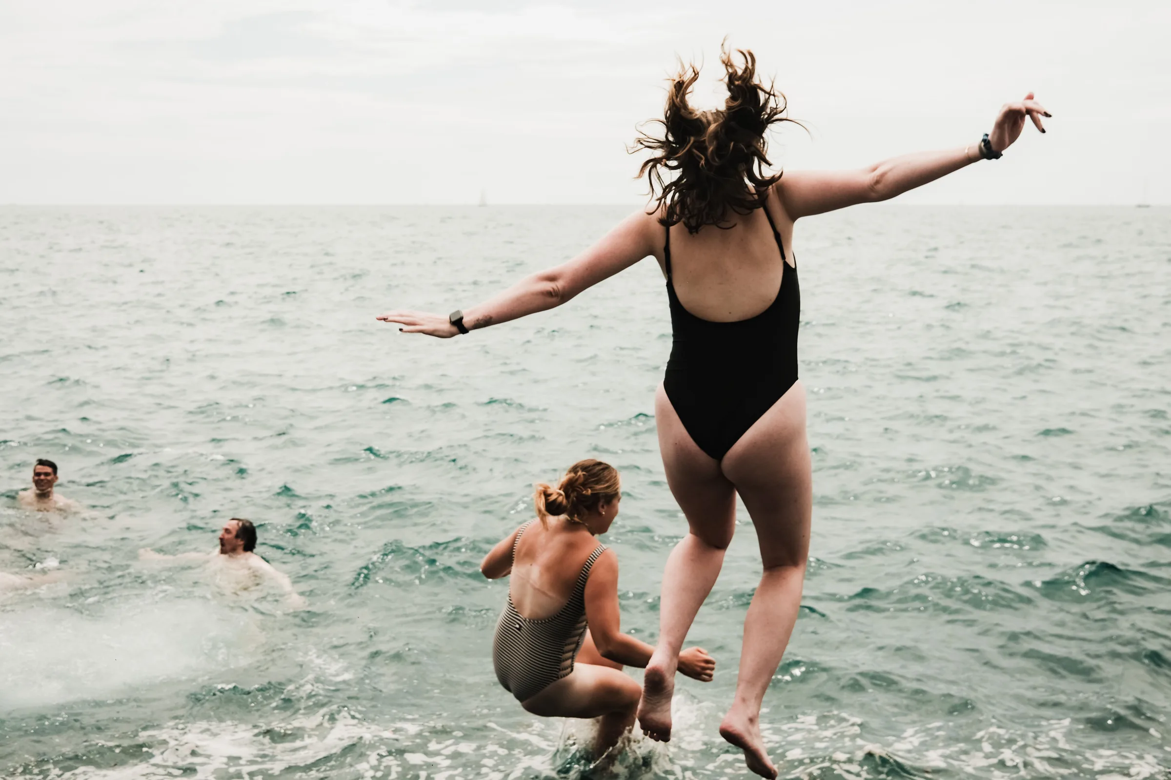 Two women leaping from the shore into a gray sea, arms flung wide