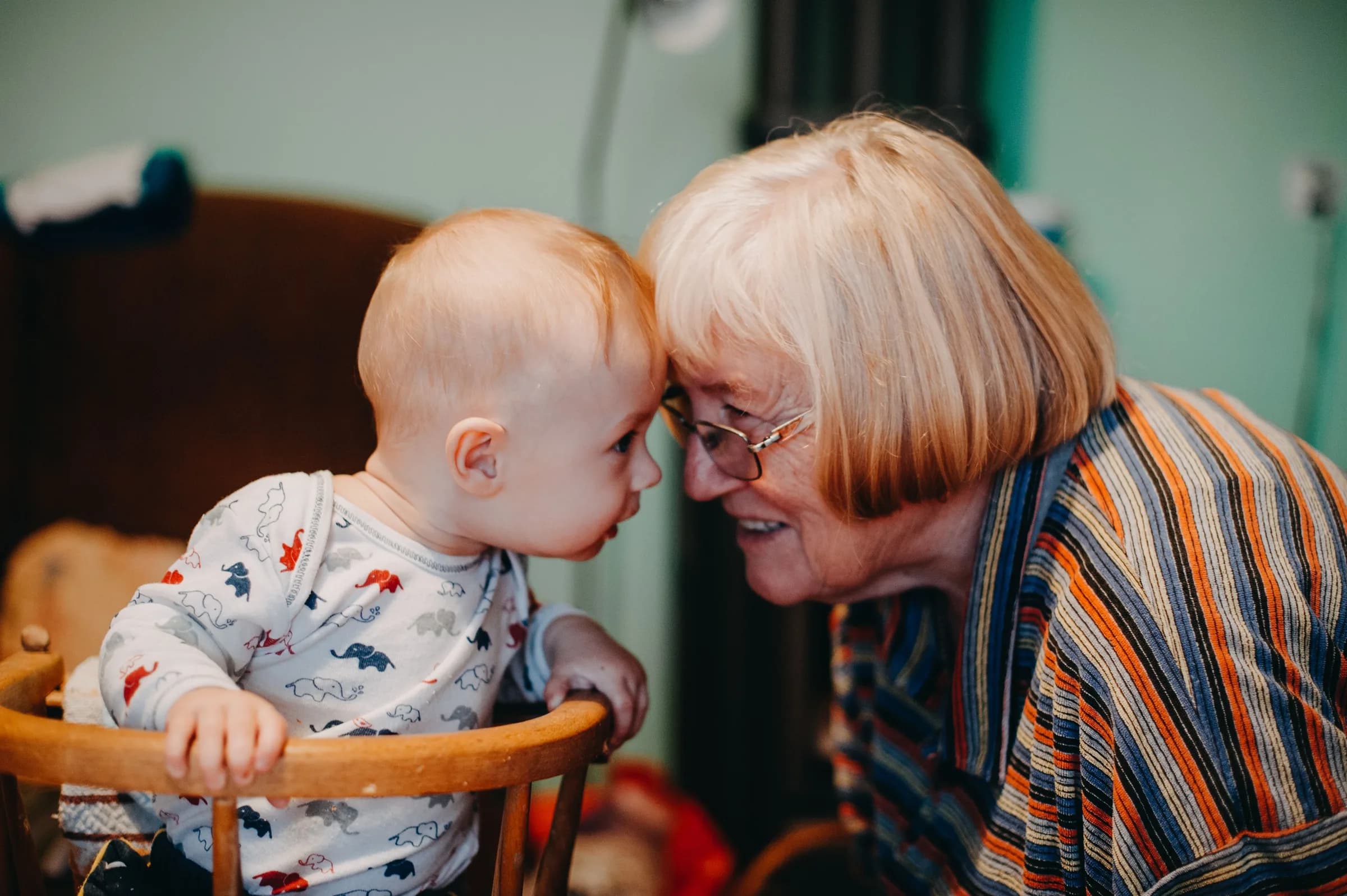 A grandmother leaning in to touch foreheads with a baby in a wooden high chair