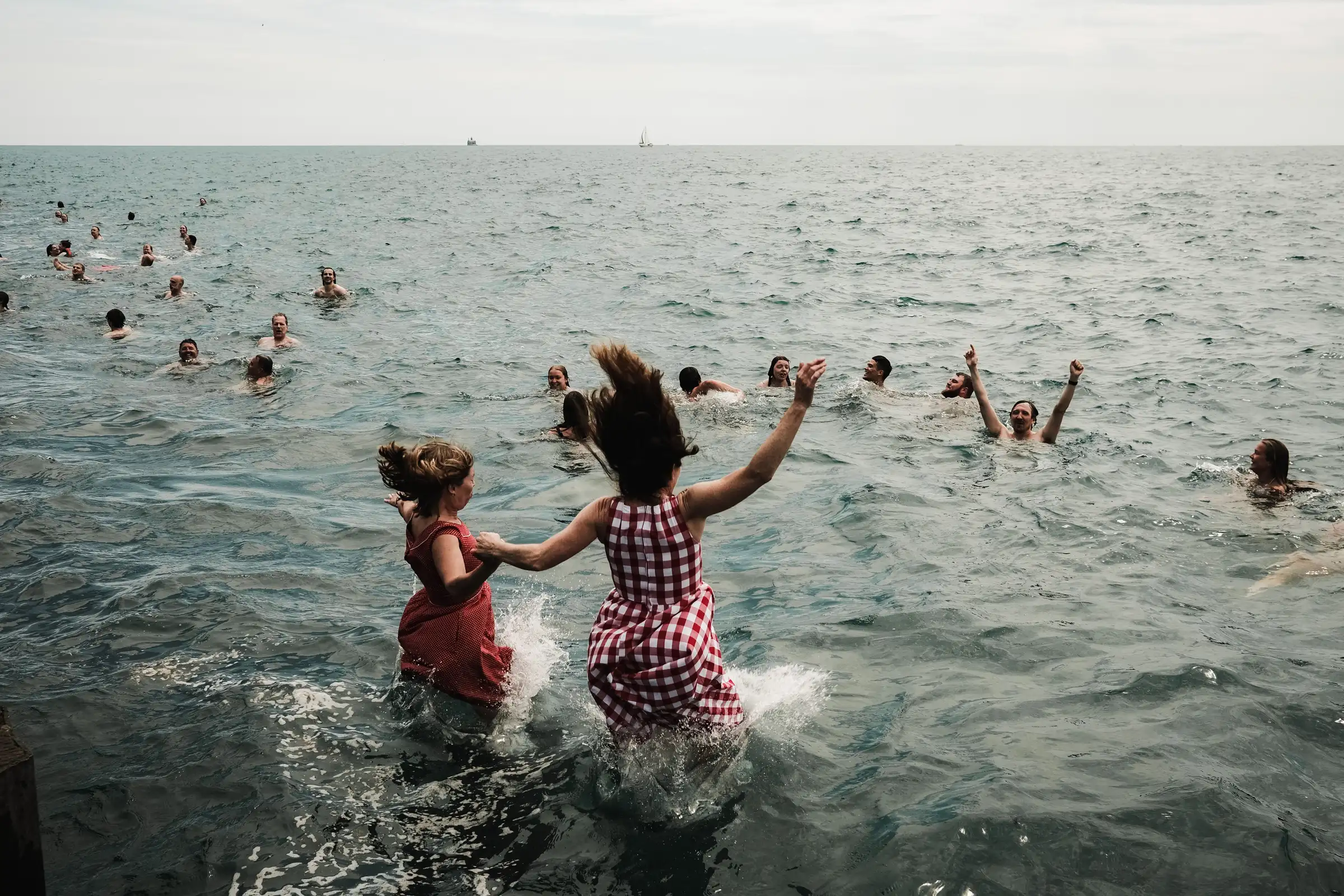 Two girls holding hands as they run splashing into the sea, swimmers scattered across the water beyond