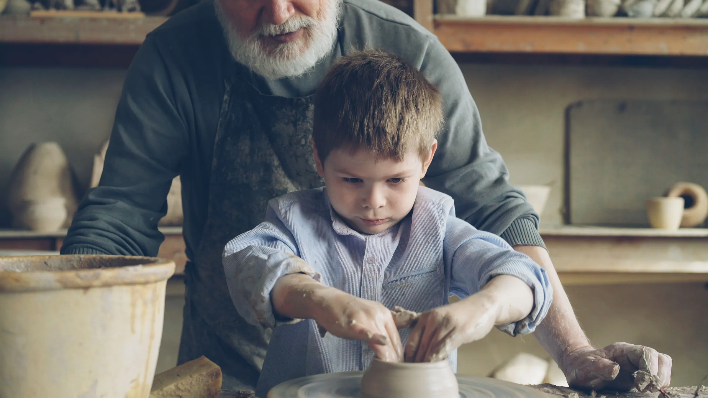 A grandfather standing behind his grandson at a potter’s wheel, guiding the boy’s clay-covered hands