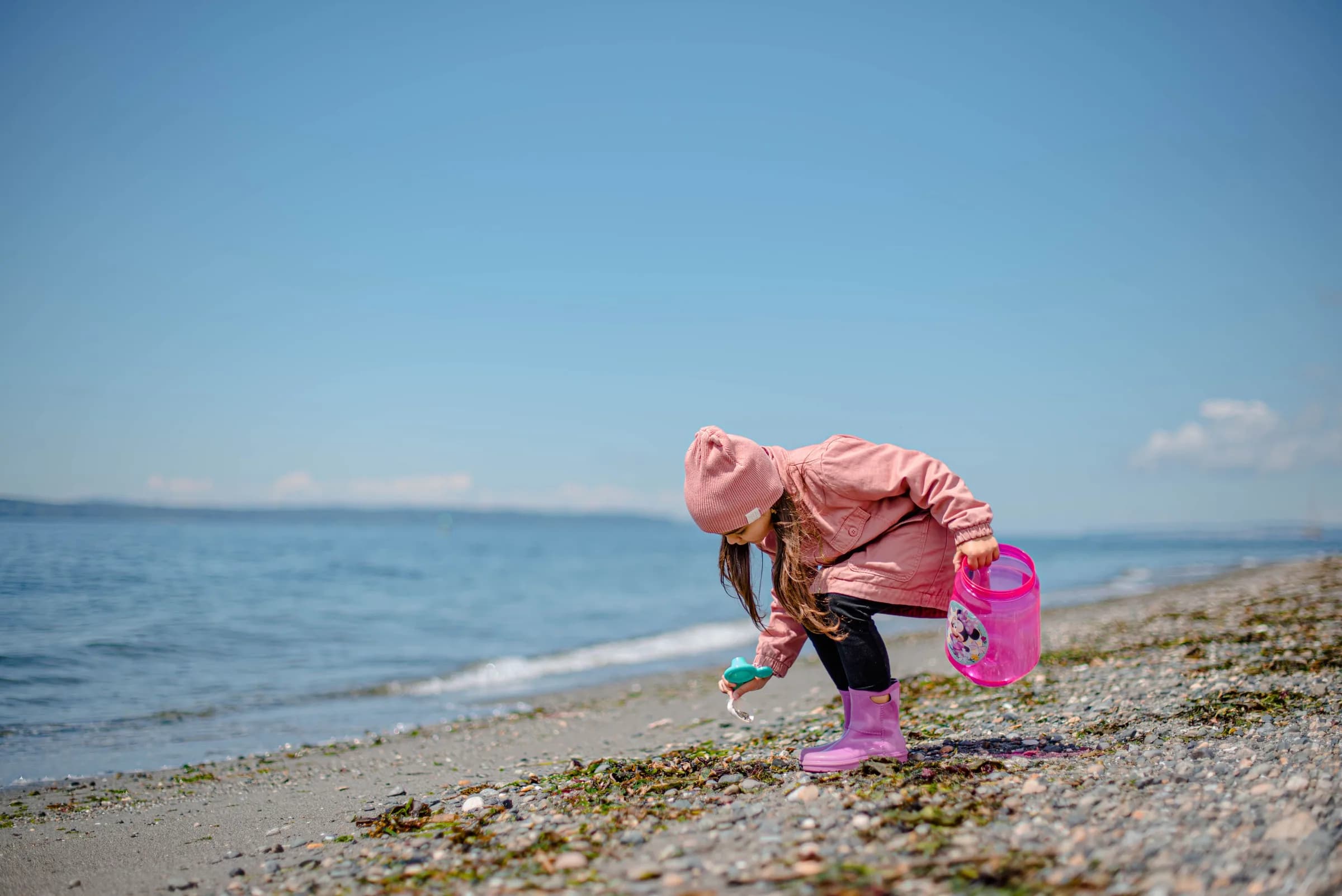 A small child in a pink coat and boots bending to pick up a shell at the edge of a wide blue sea