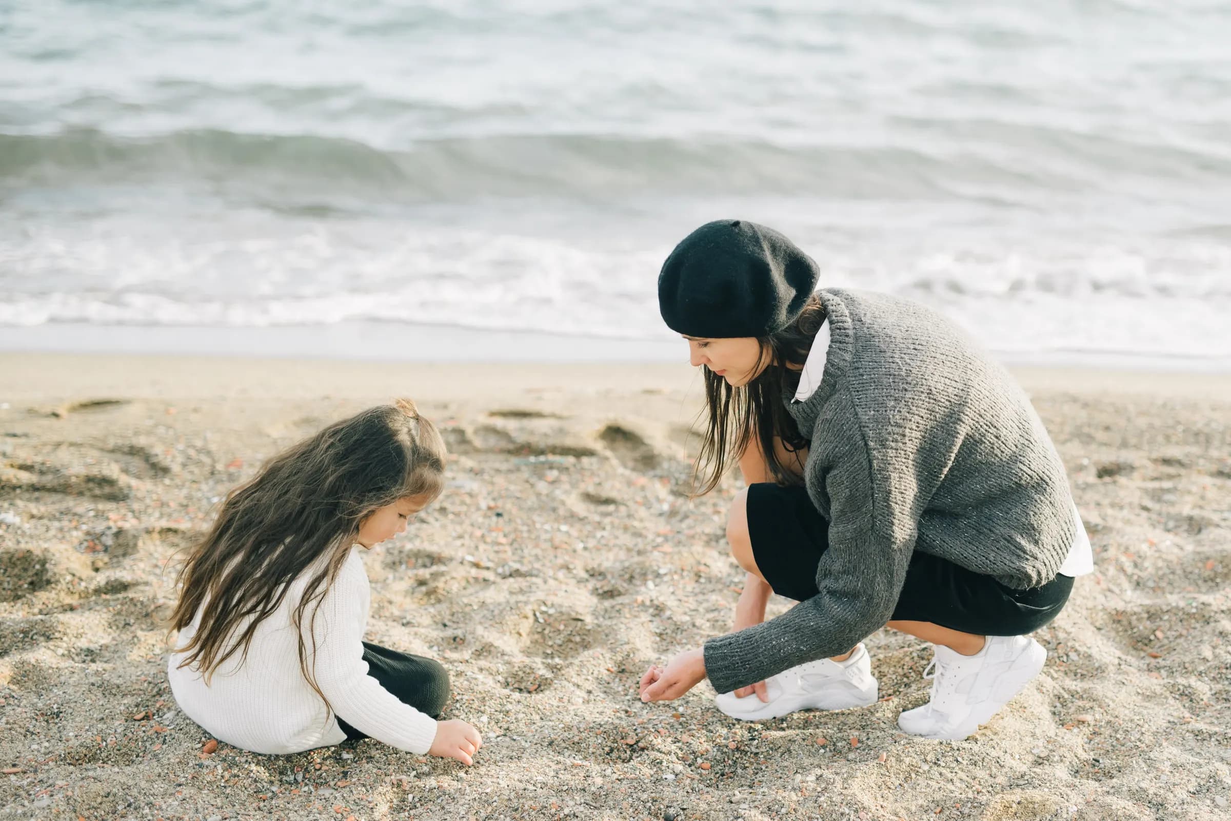 A mother and her small daughter crouched side by side on a gray beach, picking shells out of the sand