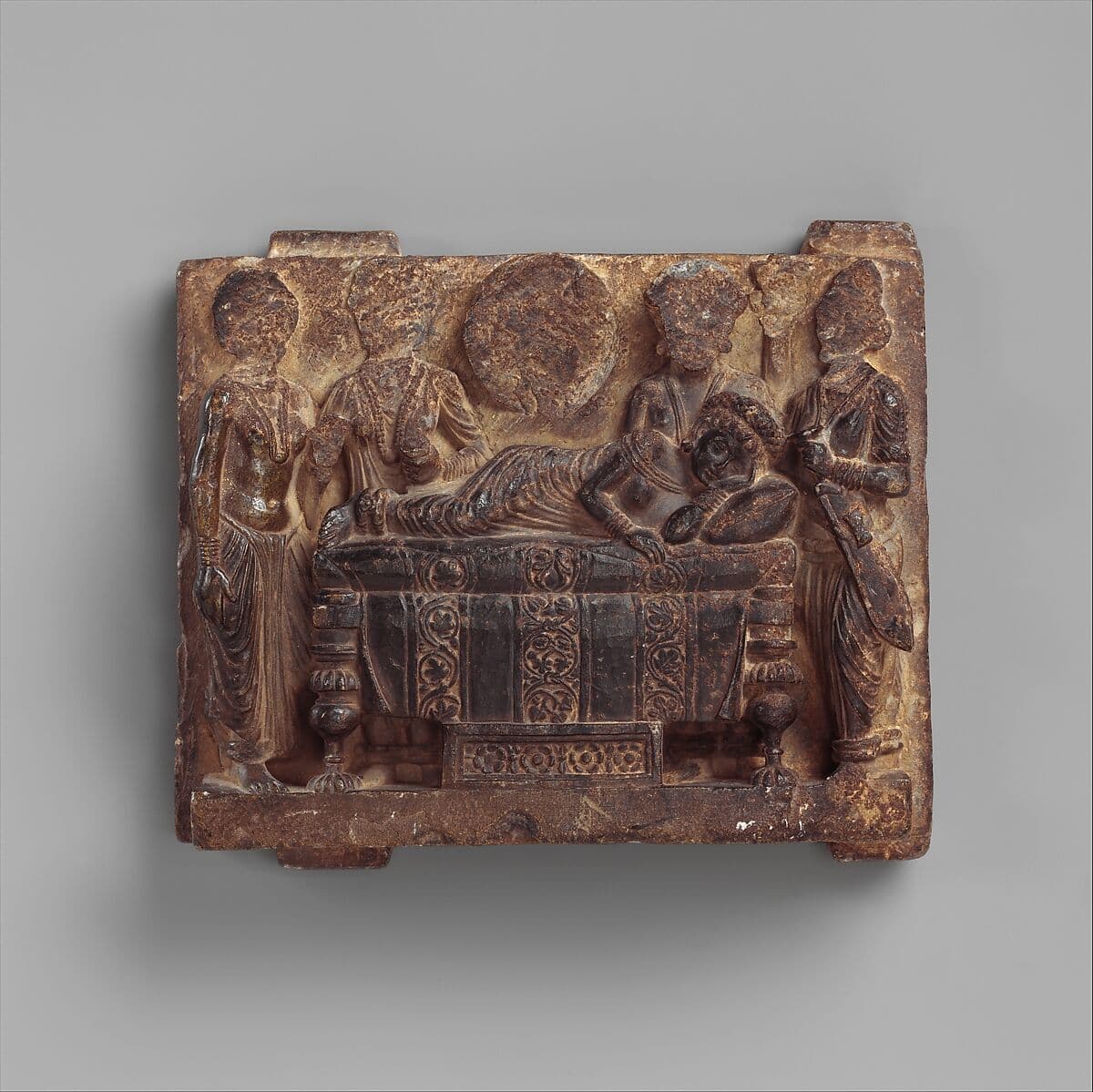 Stone relief showing Queen Maya reclining on a bed, surrounded by attendants.