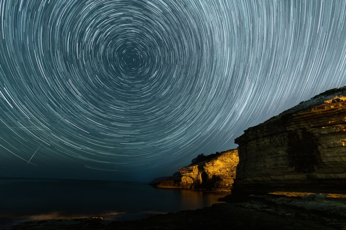 Circular star trails in the night sky above a rugged coastal cliff.