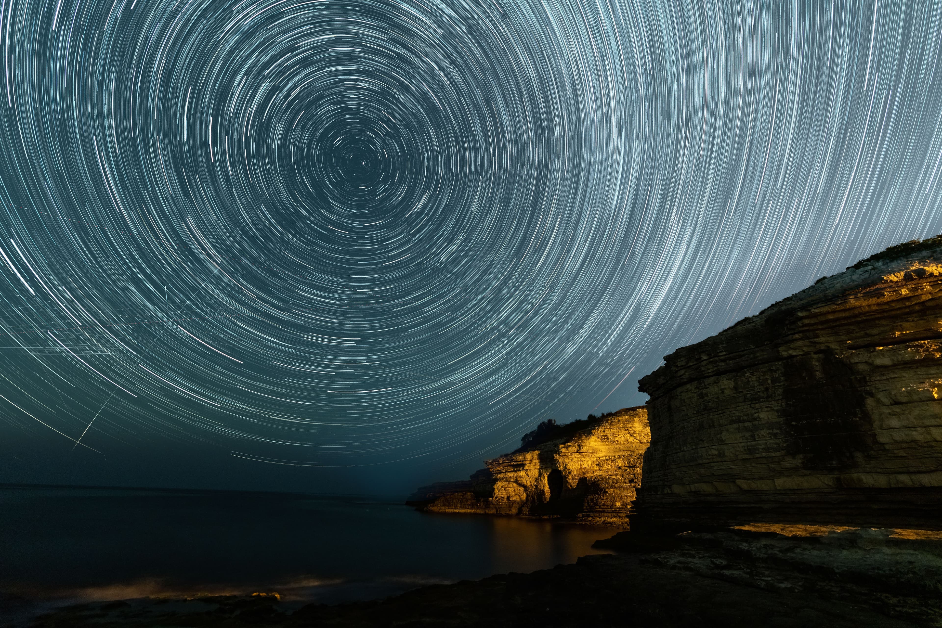 Circular star trails in the night sky above a rugged coastal cliff.