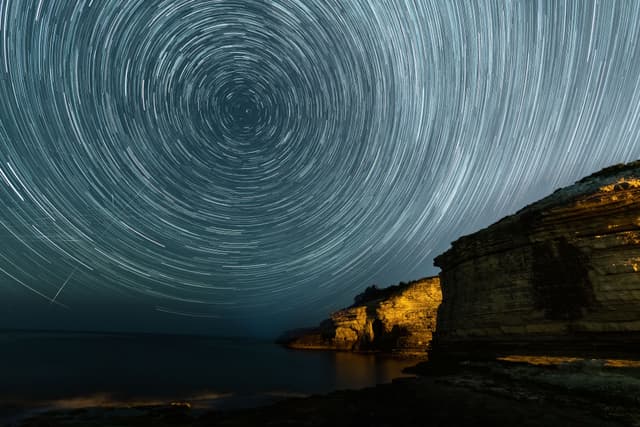 Circular star trails in the night sky above a rugged coastal cliff.