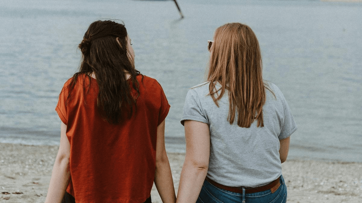 Two women holding hands beside the water, viewed from behind.