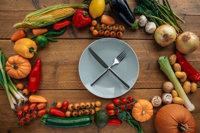 An empty ceramic plate with a knife and fork, surrounded by assorted vegetables.
