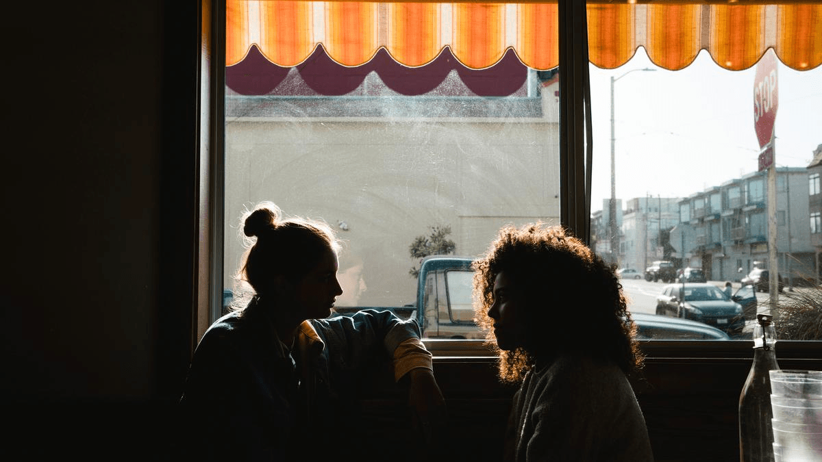 Two people talking together beside a sunlit café window.